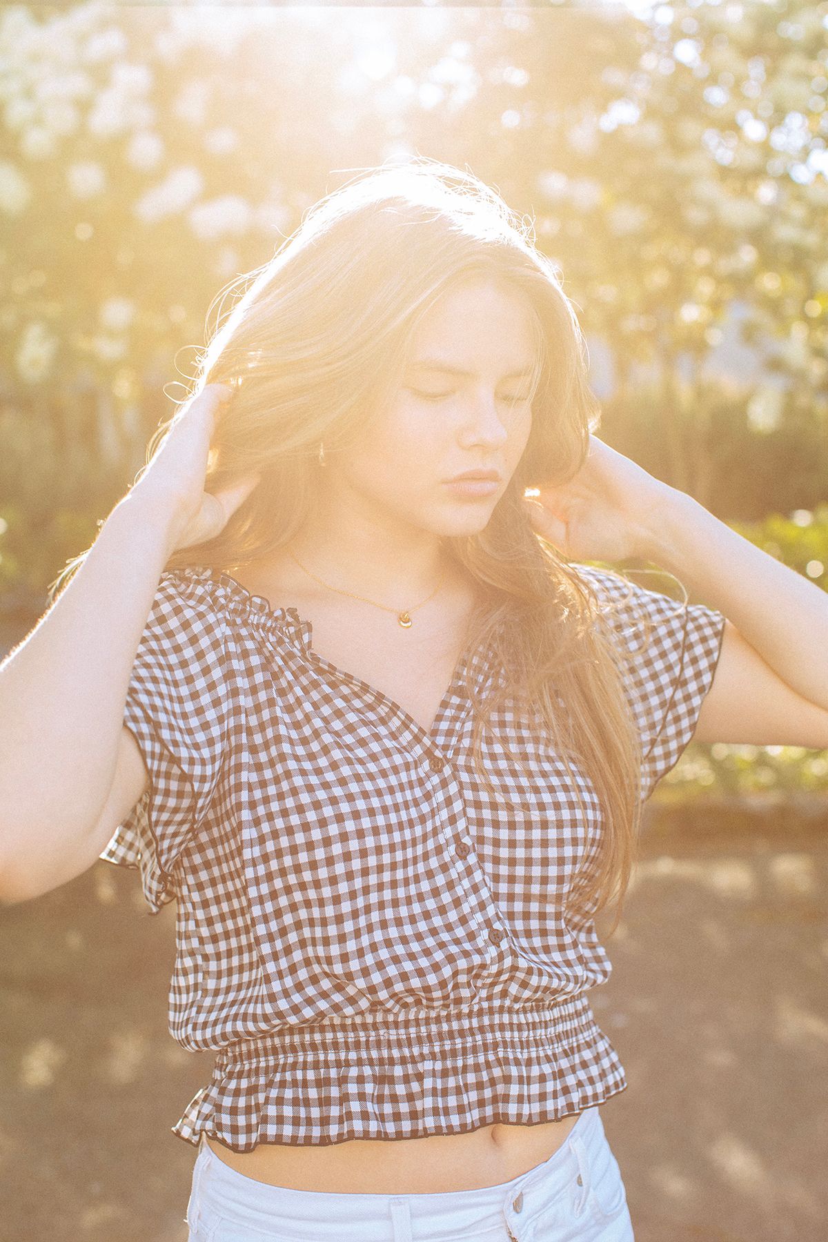 Young woman in checkered crop top poses outdoors in golden sunlight with hands in long blonde hair.