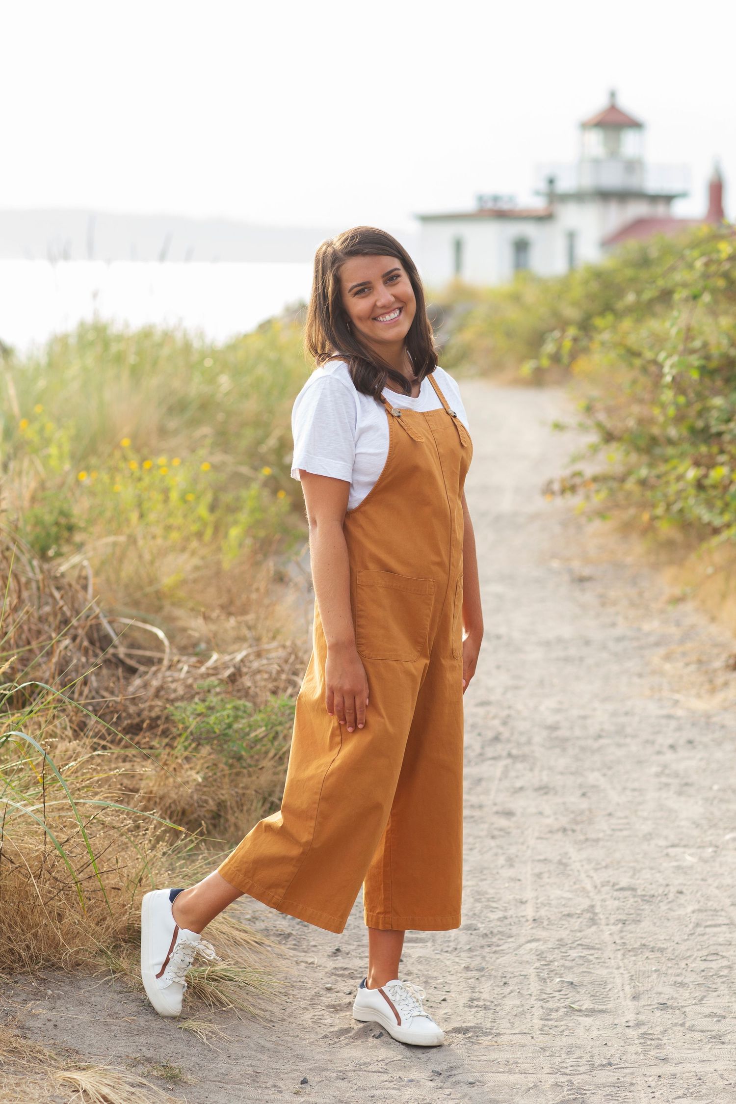 Senior Portrait Girl stands in front of lighthouse at Discovery Park with driftwood wearing orange overalls and sneakers at sunset