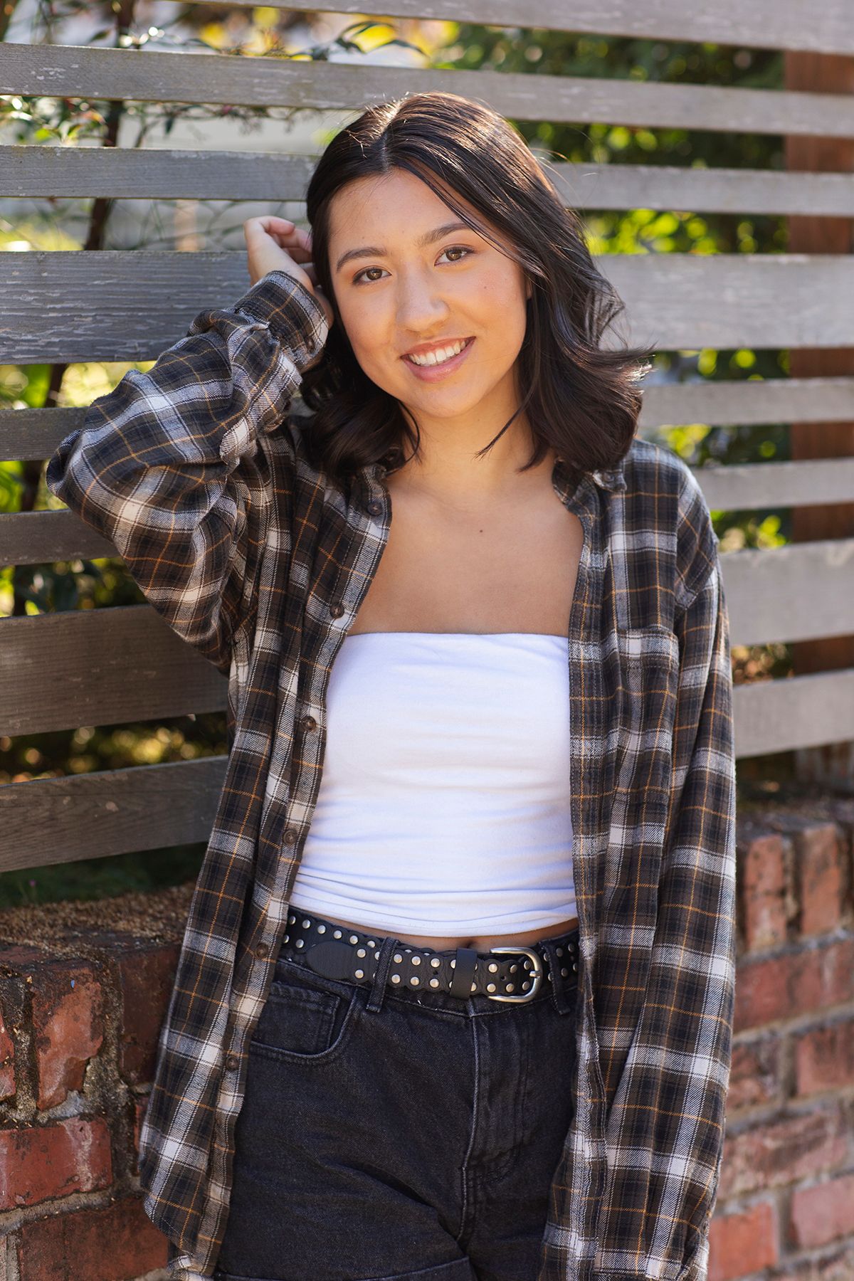 Young woman in plaid shirt and white crop top smiles outdoors by wooden fence.