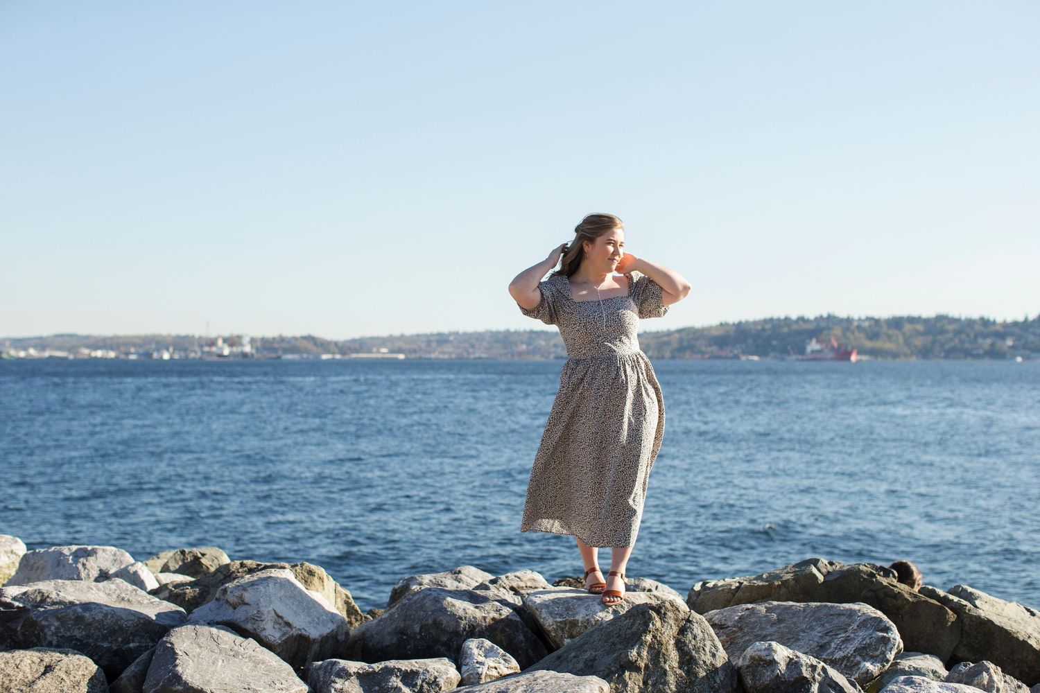 Woman in a flowy midi dress standing on rocky shoreline with blue ocean and clear sky backdrop.