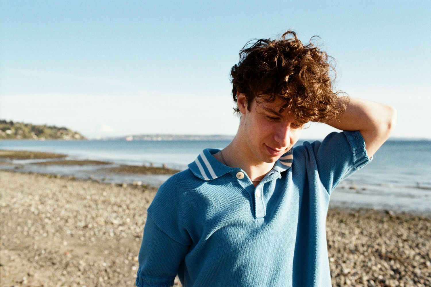 Young man with curly hair in a blue polo shirt standing on a pebble beach with ocean in the background.
