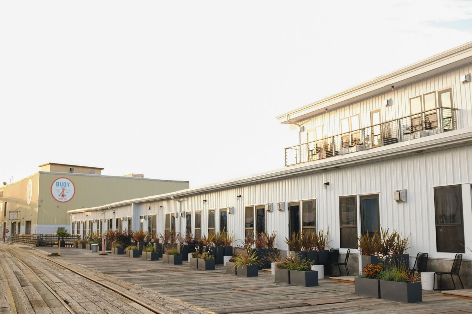 A waterfront hotel building with white siding, planters, and outdoor seating along a wooden pier boardwalk.