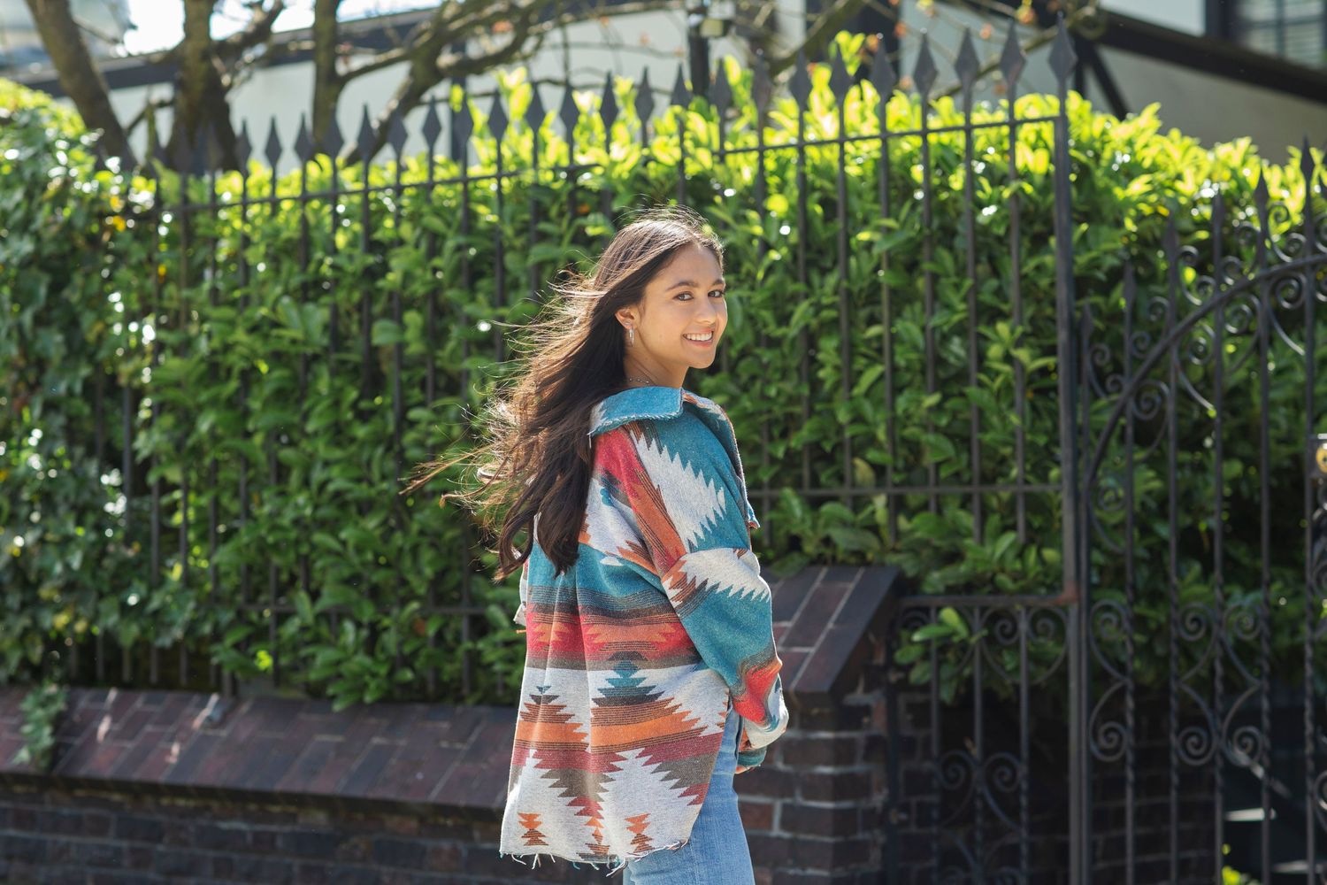 Young woman in colorful aztec print jacket smiling over her shoulder on a sunny street.