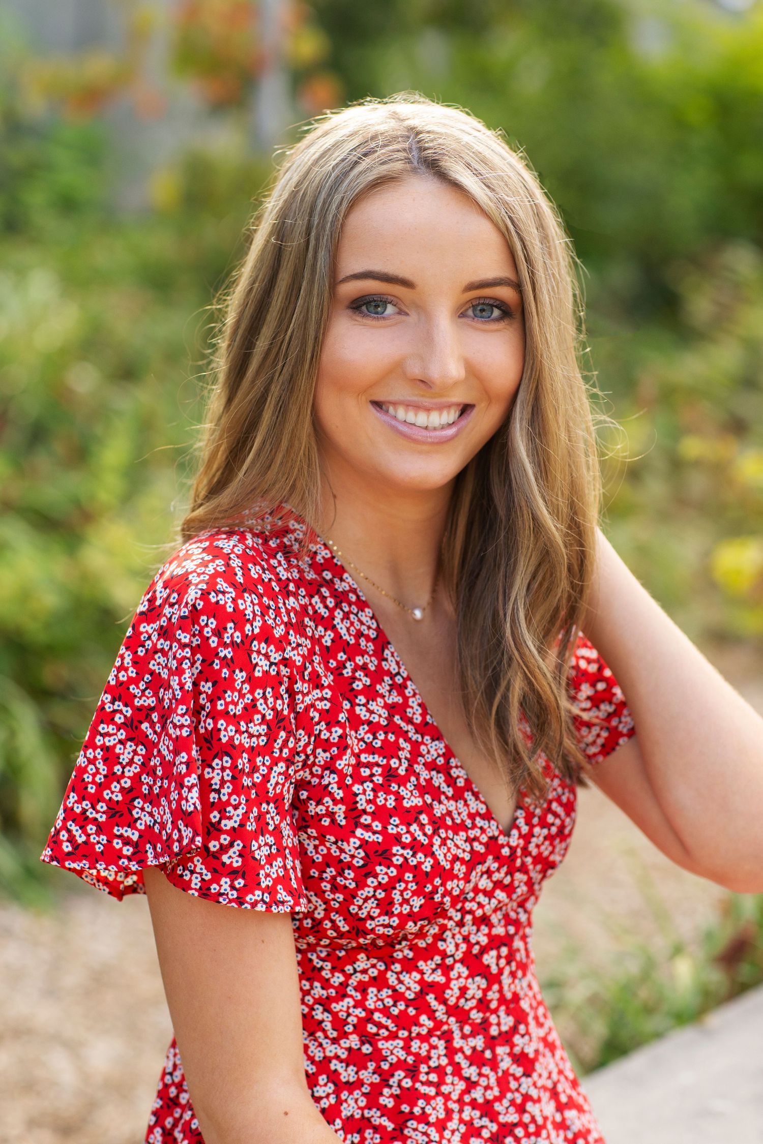 Smiling blonde woman with blue eyes in red floral dress in a garden setting.