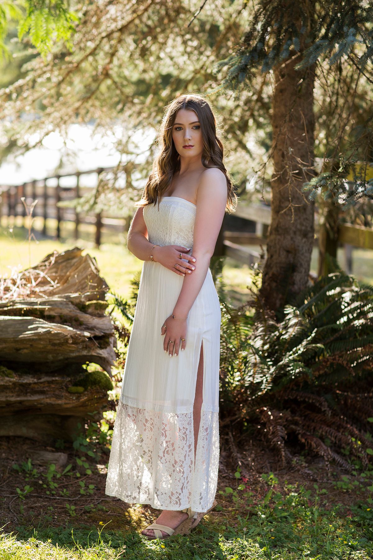 Young woman in white strapless lace maxi dress posing outdoors near pine trees and rustic wooden fence.