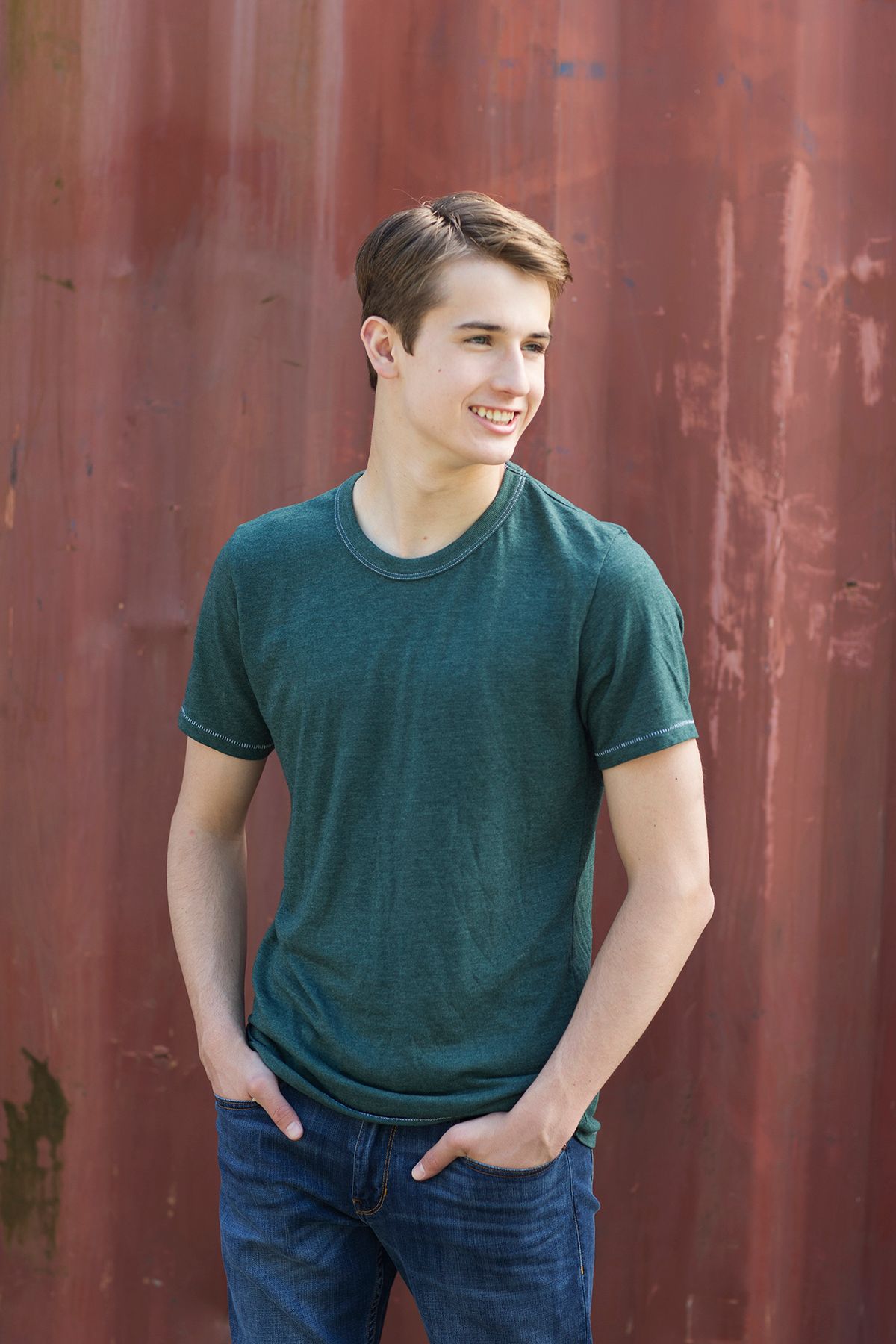 Smiling young man in green t-shirt and jeans standing against a rusty red metal wall.