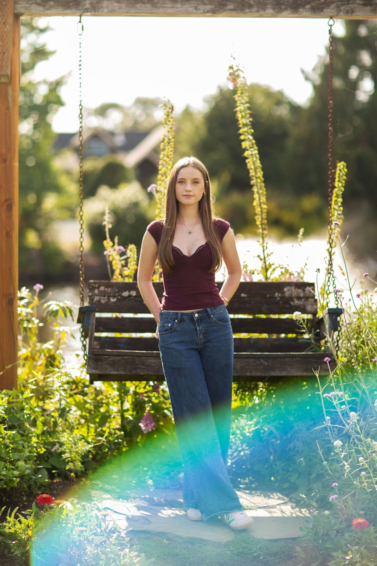 Young woman in burgundy top and jeans stands before a wooden garden swing surrounded by blooming flowers in sunlight.