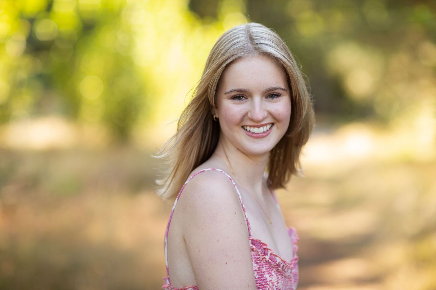 Smiling blonde teenage girl in pink dress posing on a sunlit outdoor path.