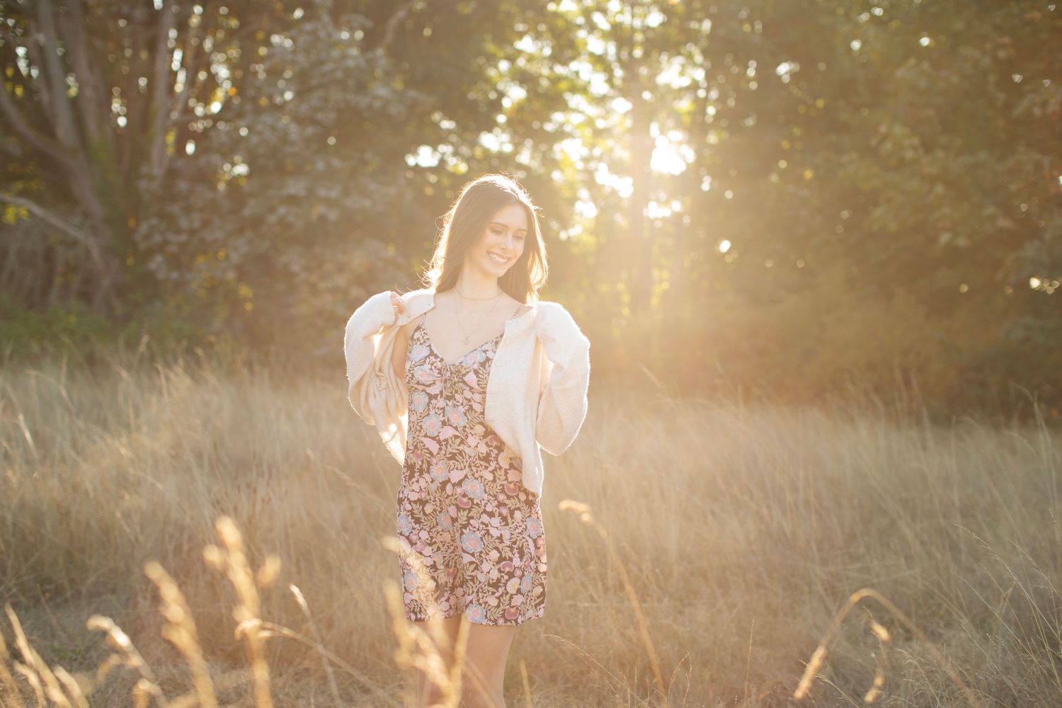 Young woman in floral dress and white cardigan standing in golden sunlit field.