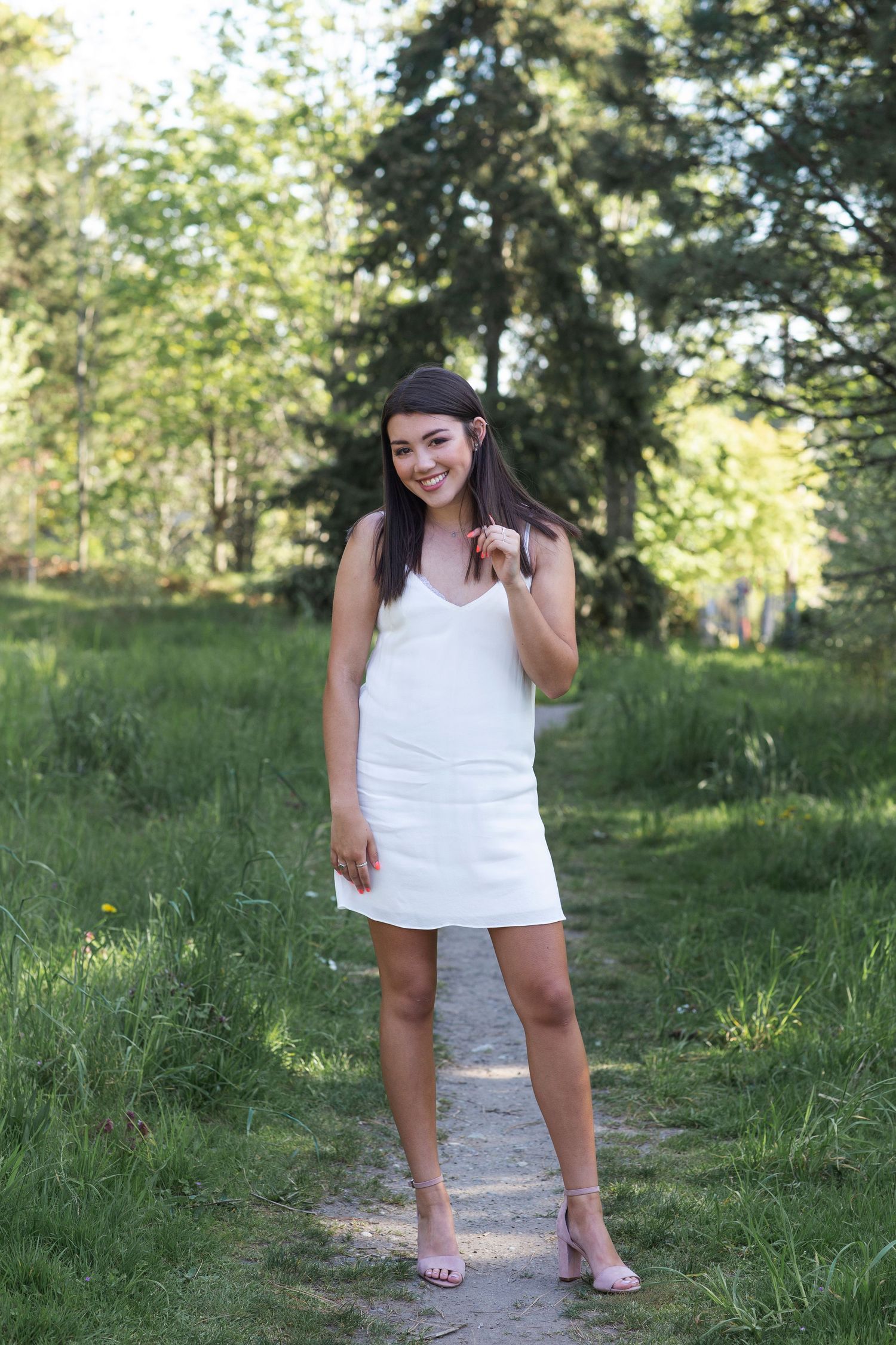 Young woman in white mini dress and heels smiling on a garden path surrounded by lush green trees.