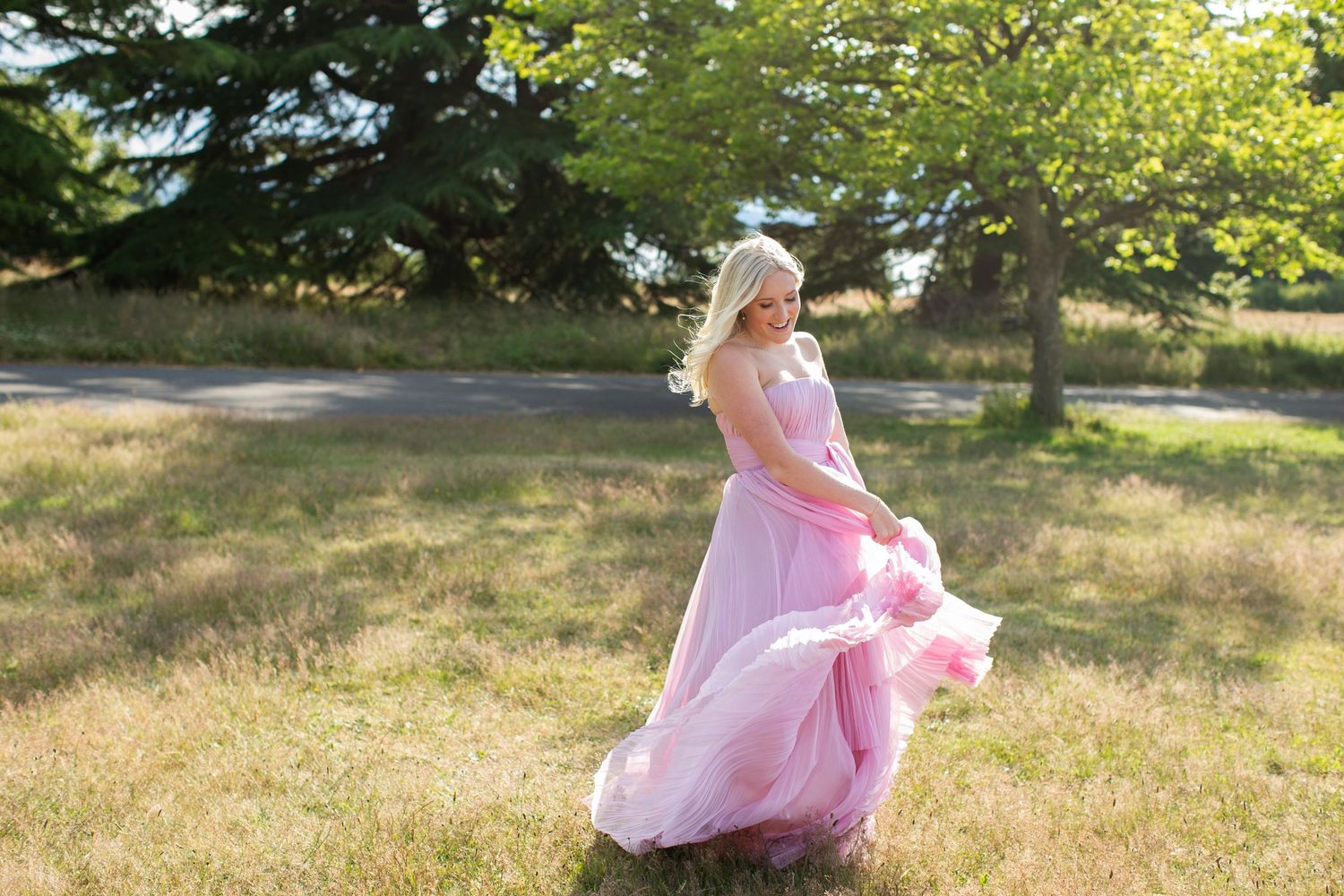 Blonde woman twirling in a flowing pink tulle gown in an open grassy field with trees behind her.