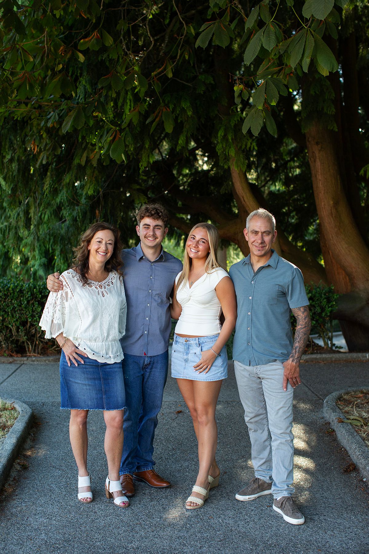 Family of four smiling together outdoors under lush green trees on a sunny day.