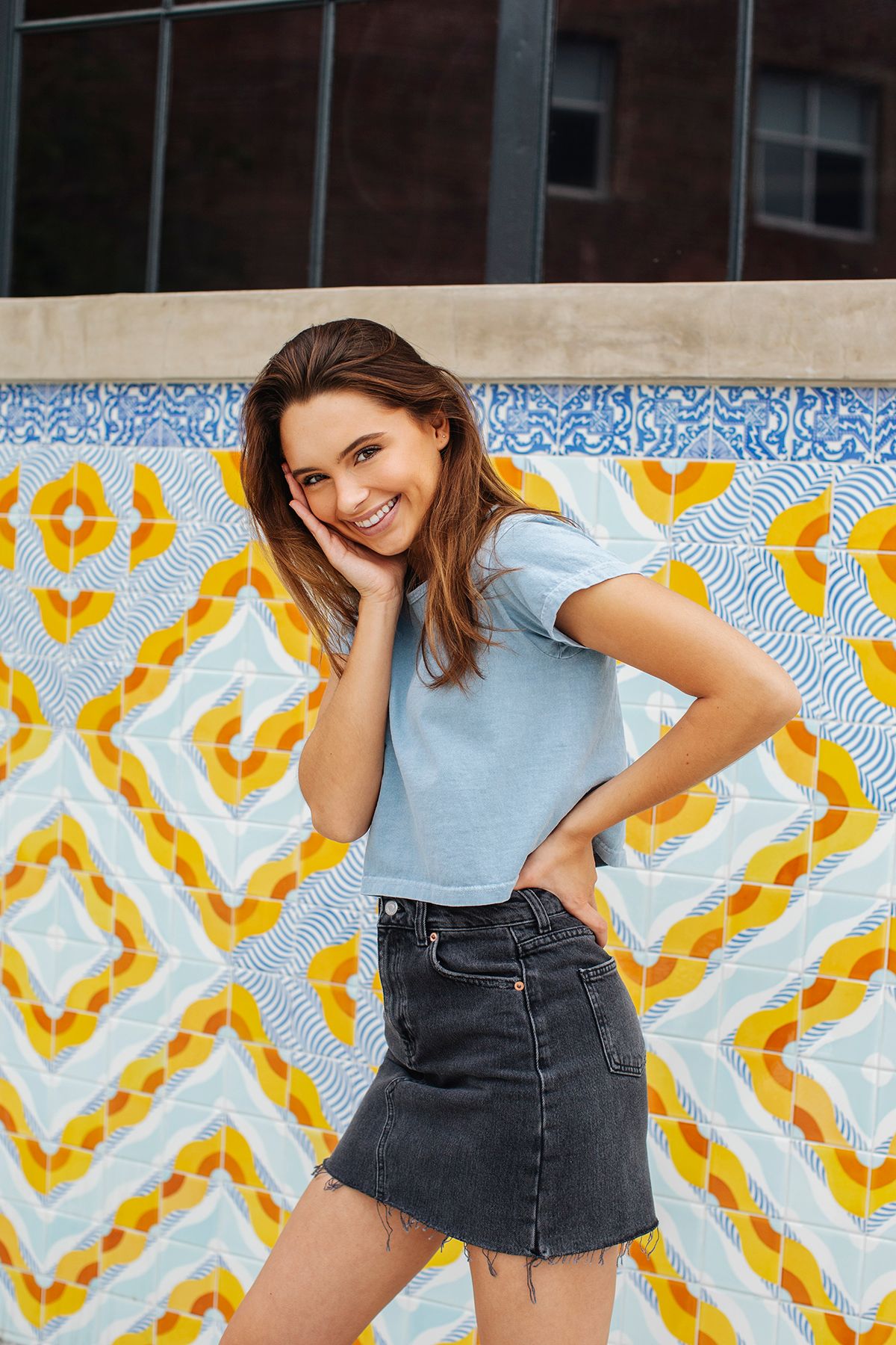 Smiling young woman in blue crop top and denim skirt posing against colorful geometric tile wall.