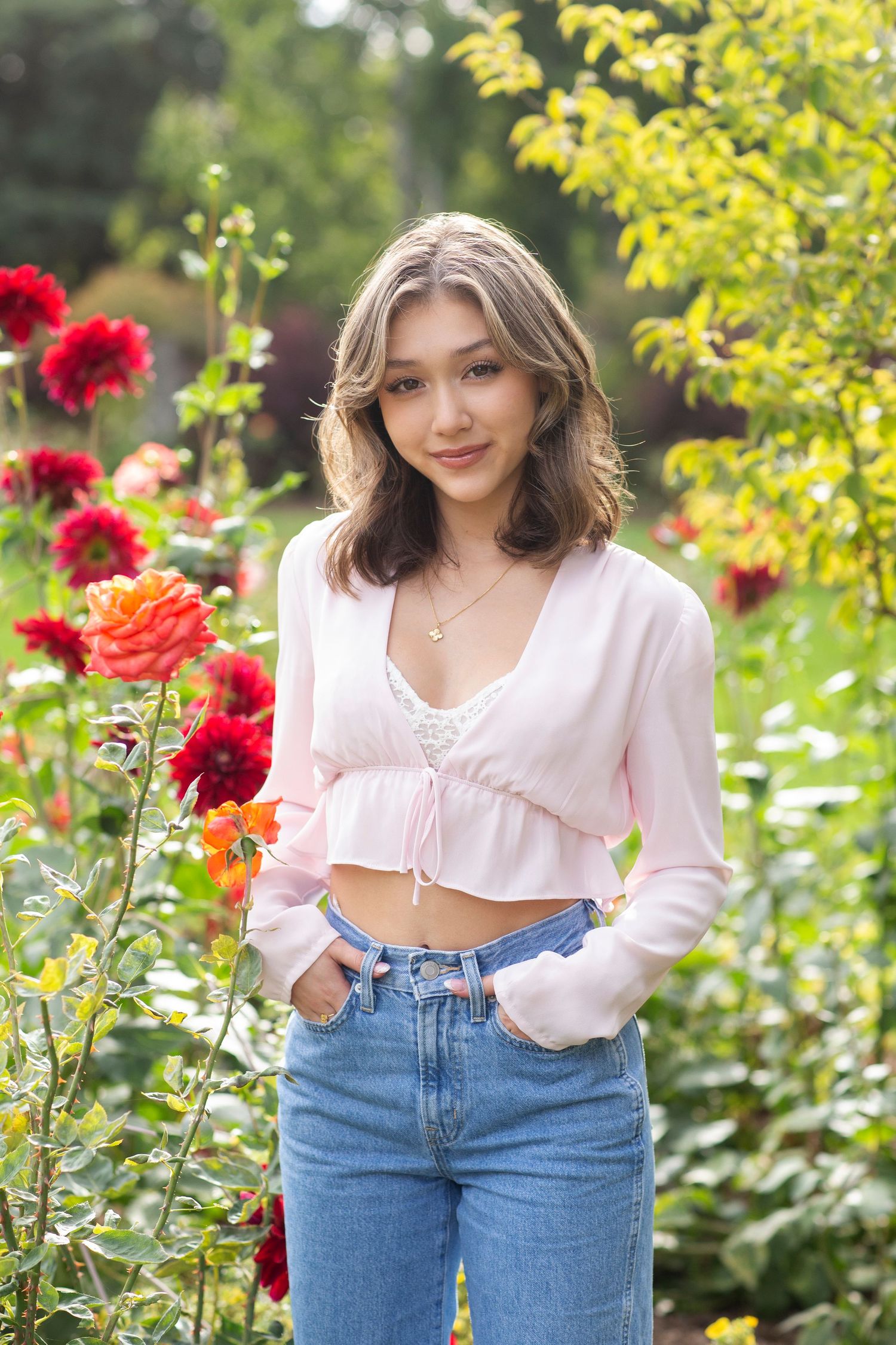 Teen girl in pink cardigan and jeans posing among vibrant red and orange dahlias in a garden.