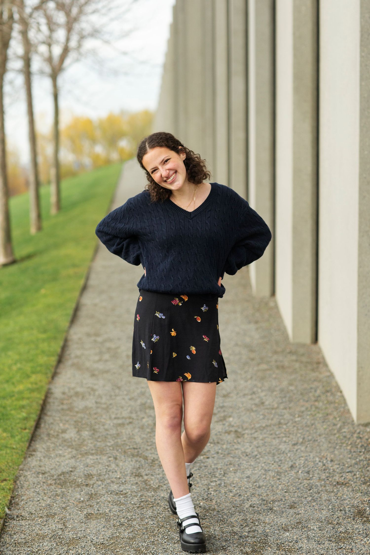 Young woman smiling in navy sweater and floral skirt walking along a modern building pathway in autumn.
