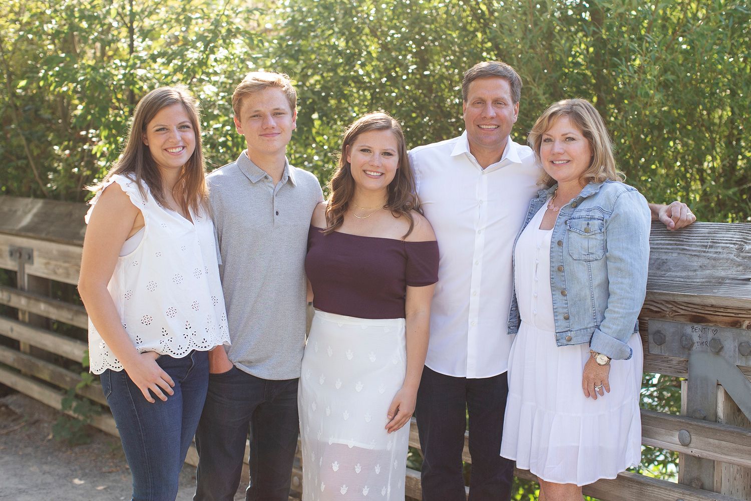 A smiling family of five poses together on a wooden bridge surrounded by lush green trees.