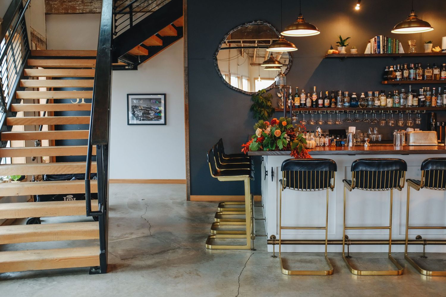 Modern bar interior with staircase, dark blue accent wall, round mirror, pendant lights, and gold bar stools.