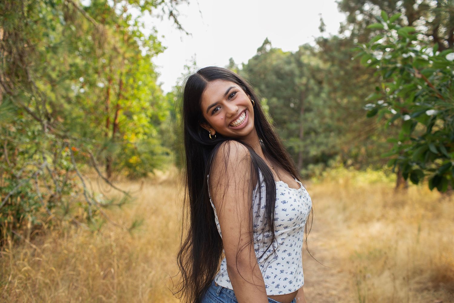 Smiling young woman in floral tank top standing on dry grass path surrounded by trees.