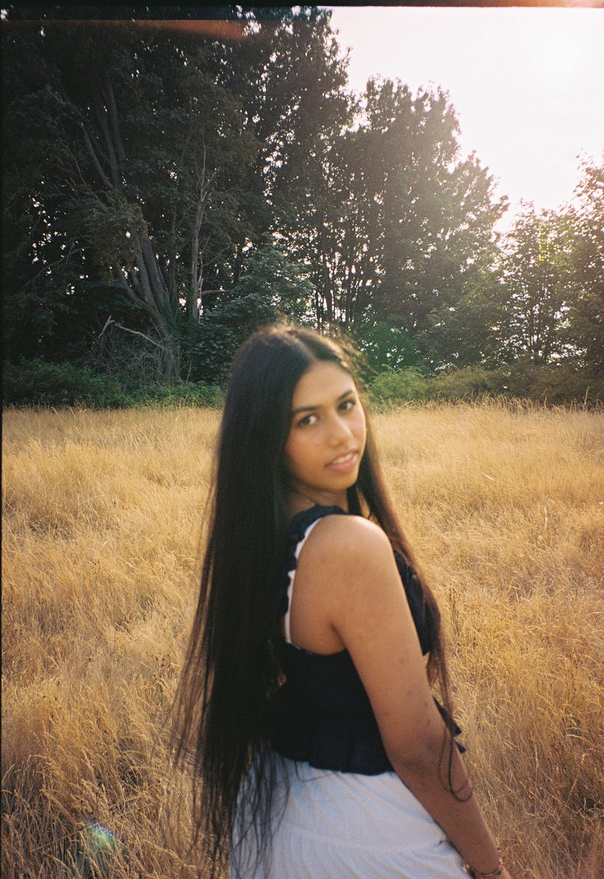 Young woman with long dark hair in black top looking back over shoulder in golden field.