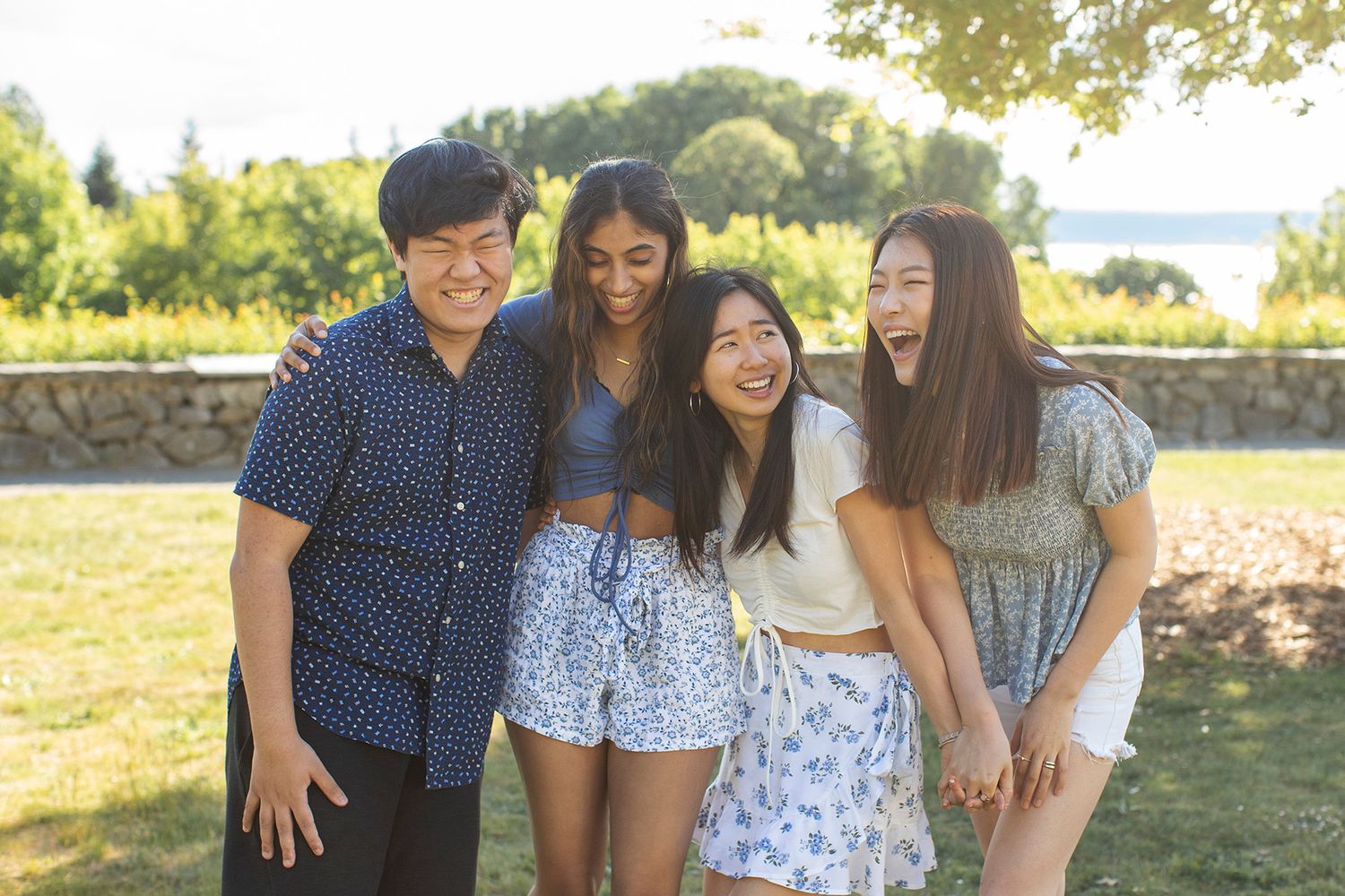 Four young friends laughing and hugging outdoors in a sunny park setting.