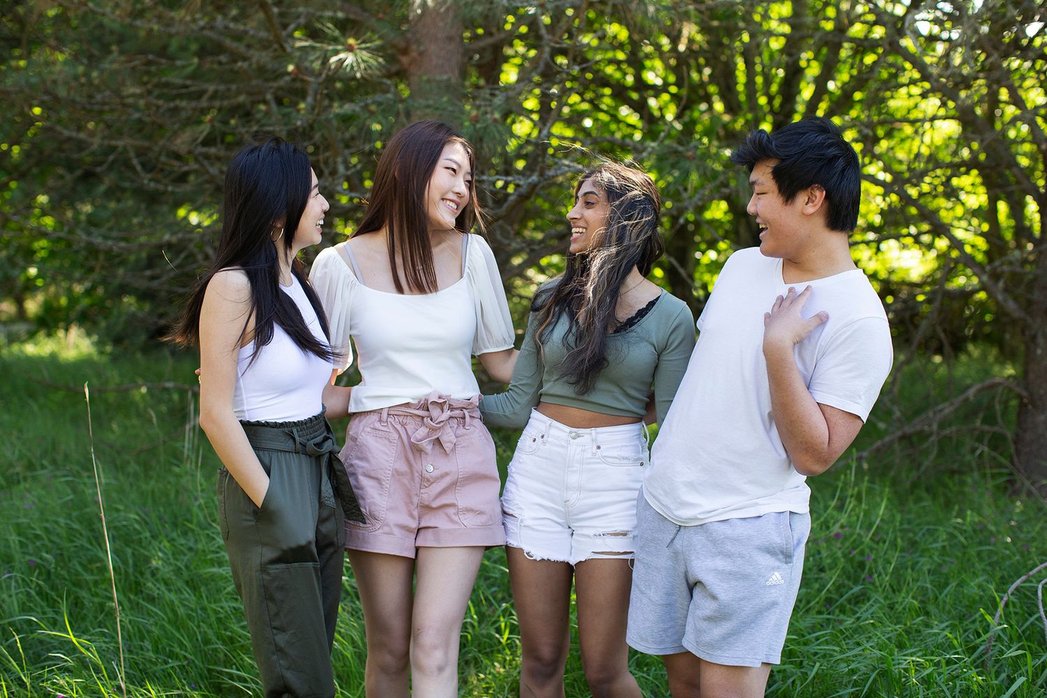 Four young friends laughing and talking together outdoors in a lush green park on a sunny day.