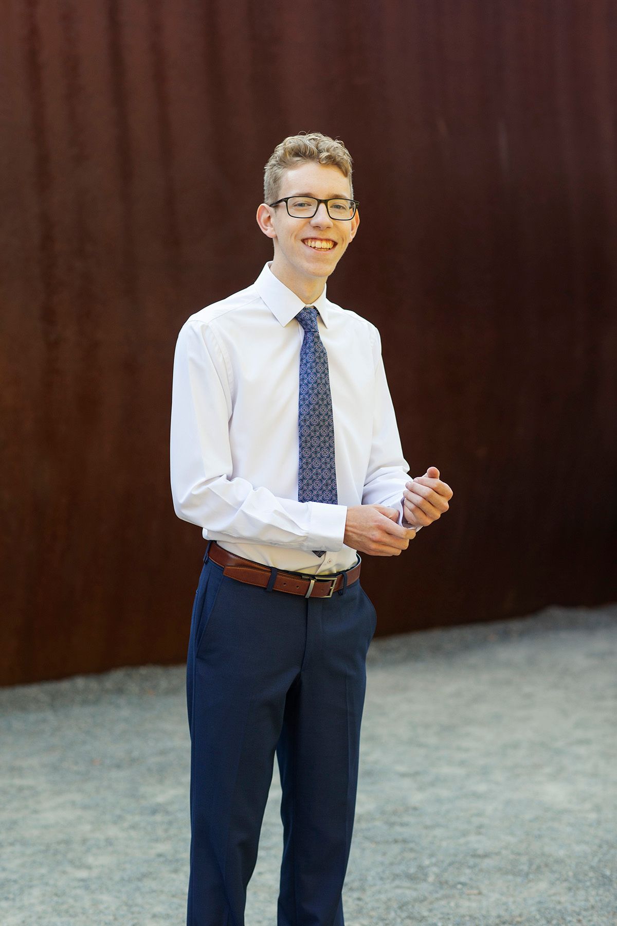 Smiling teenage boy in white dress shirt, blue patterned tie, and navy pants standing outdoors against a rustic metal wall.