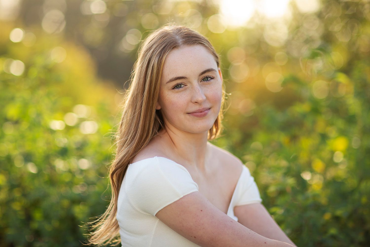 Young woman with long blonde hair smiling outdoors in a sunlit green field, wearing a white off-shoulder top.