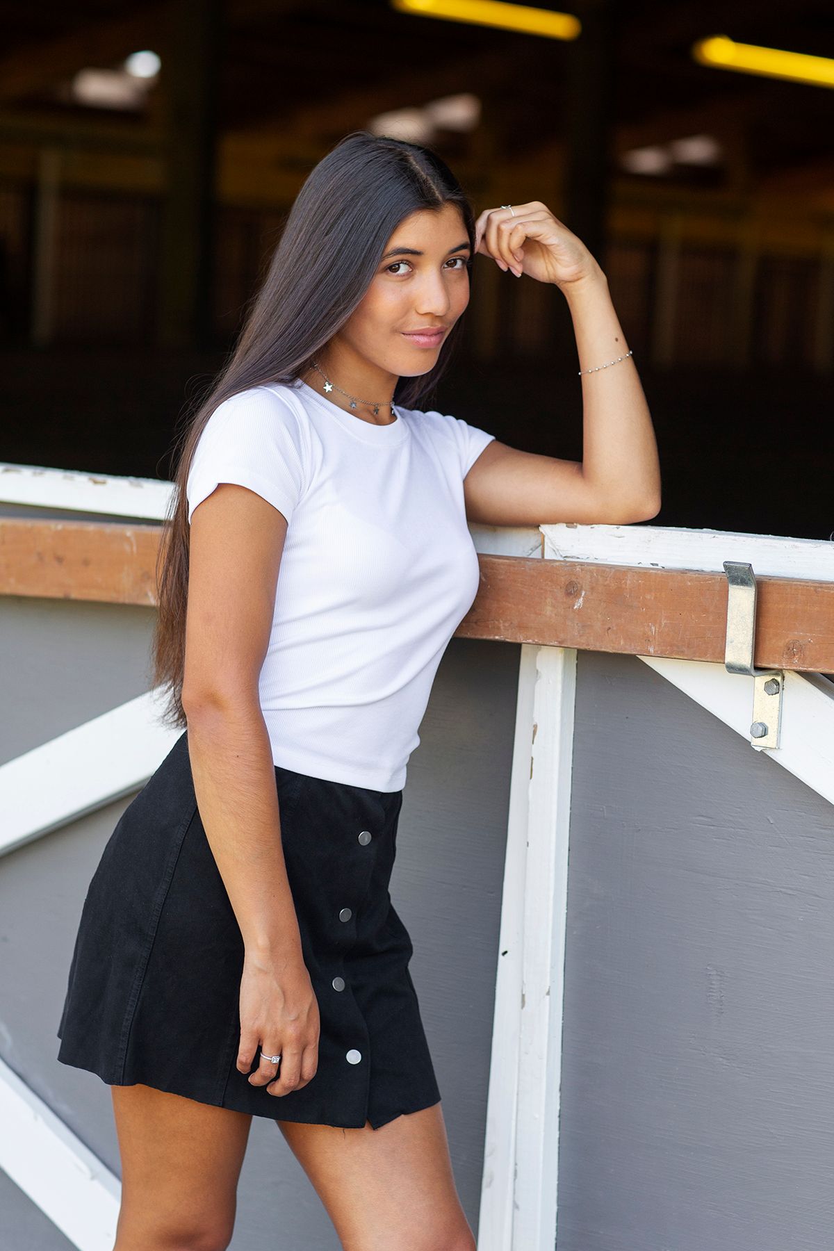 Young woman in white t-shirt and black mini skirt leaning on railing, posing outdoors with long dark hair.