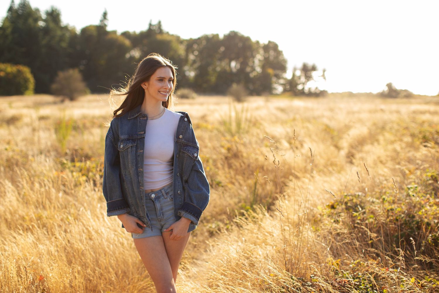 Smiling young woman in denim jacket and shorts posing in sunny golden grass field.