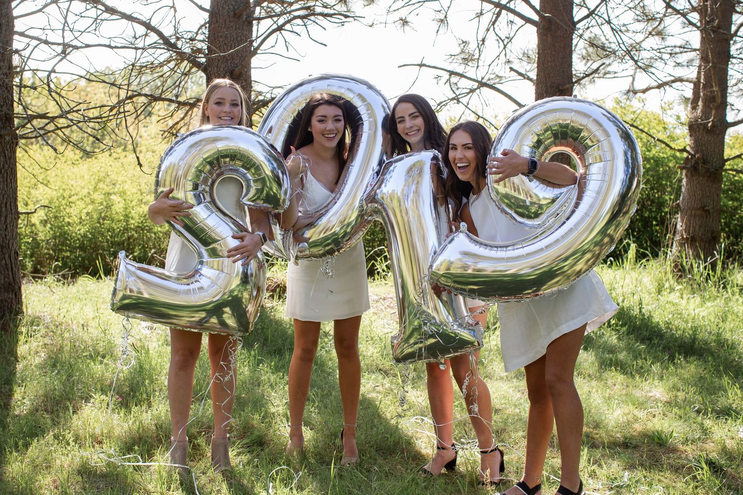 Four girls in white dresses holding silver balloon numbers 3219 outdoors among trees.