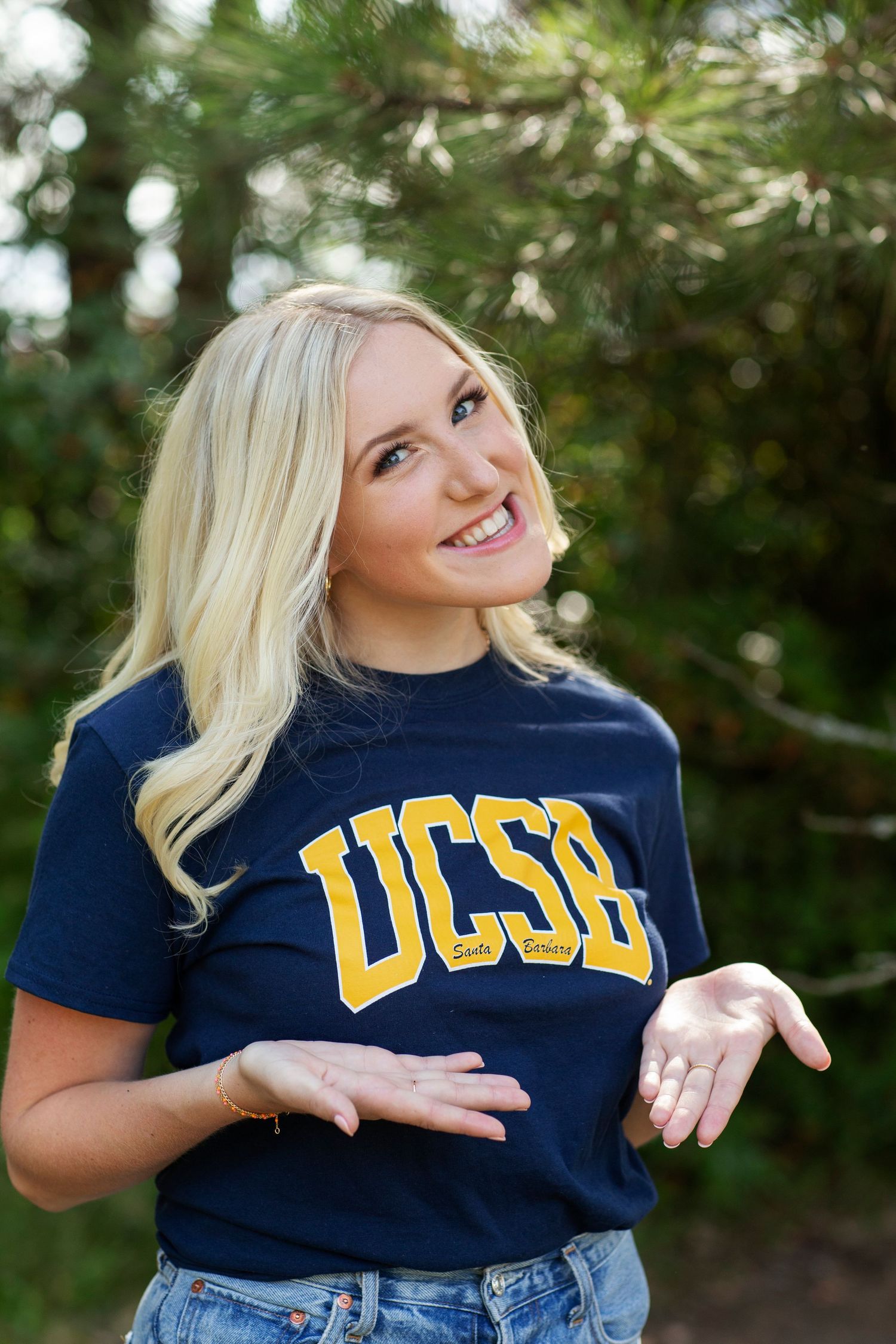Blonde woman in a navy UCSB t-shirt gesturing with hands and smiling in front of green trees.