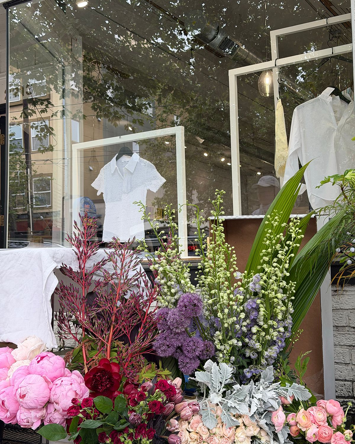 Boutique storefront display with white shirts and lush floral arrangement featuring pink peonies, purple blooms, and greenery.
