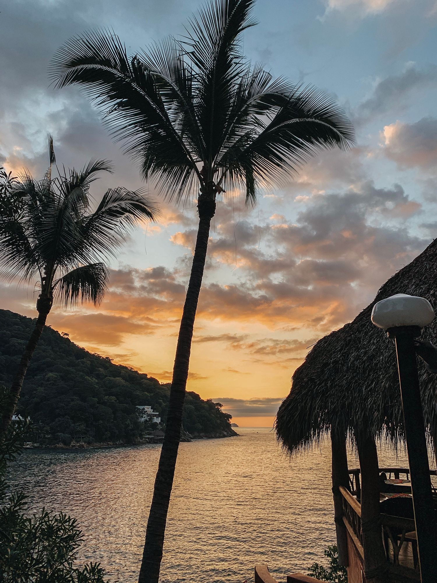 Stunning tropical sunset over calm ocean bay with tall palm trees and thatched palapa roof in Mexico.