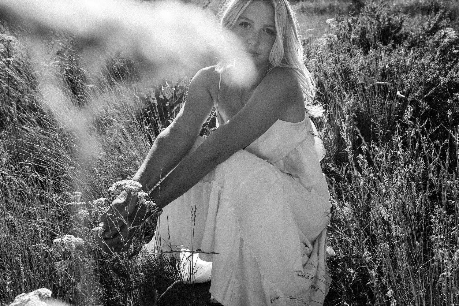 Black and white photo of a blonde woman in a white dress sitting among wildflowers in a sunny field.