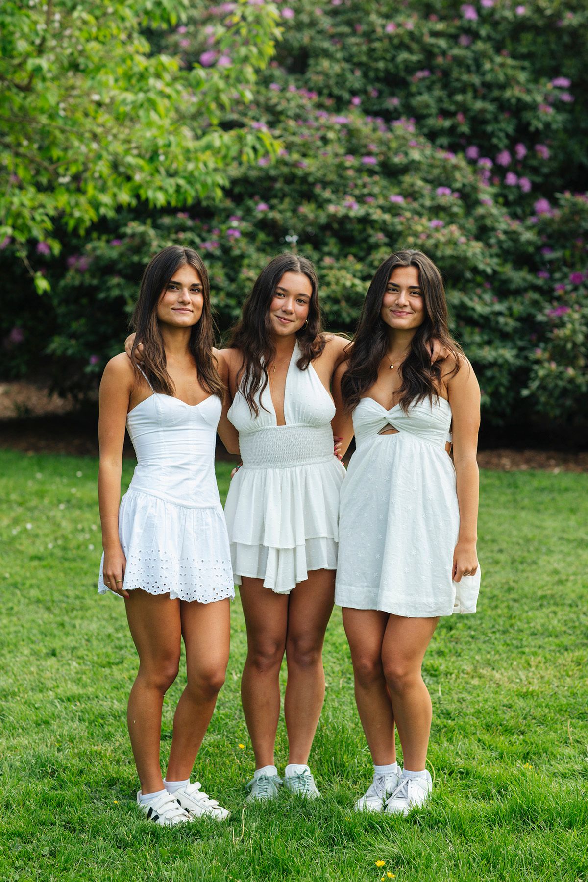 Three young women in white dresses pose together on green grass with blooming purple bushes in the background.