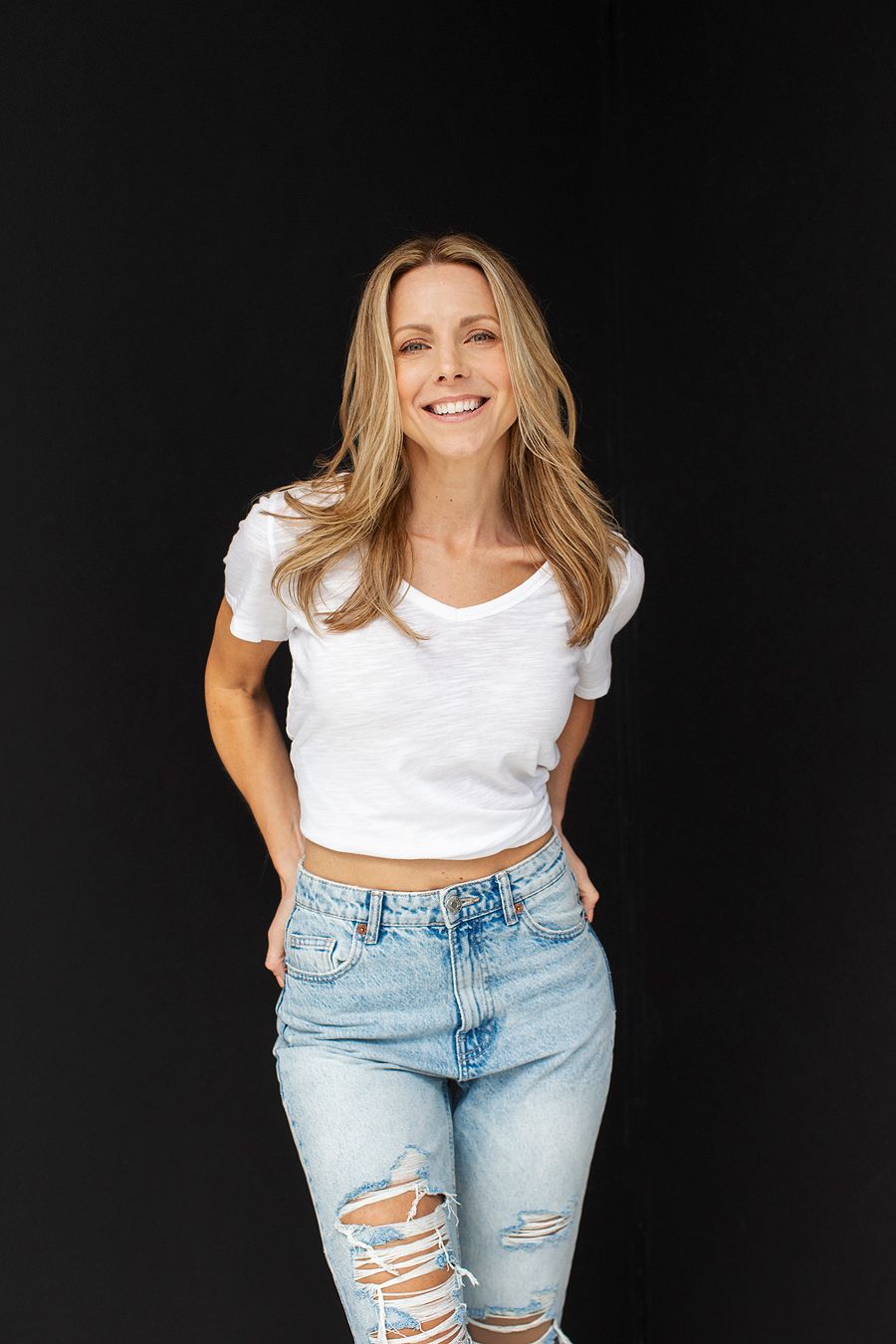 Smiling blonde woman in white crop top and ripped jeans on black background.