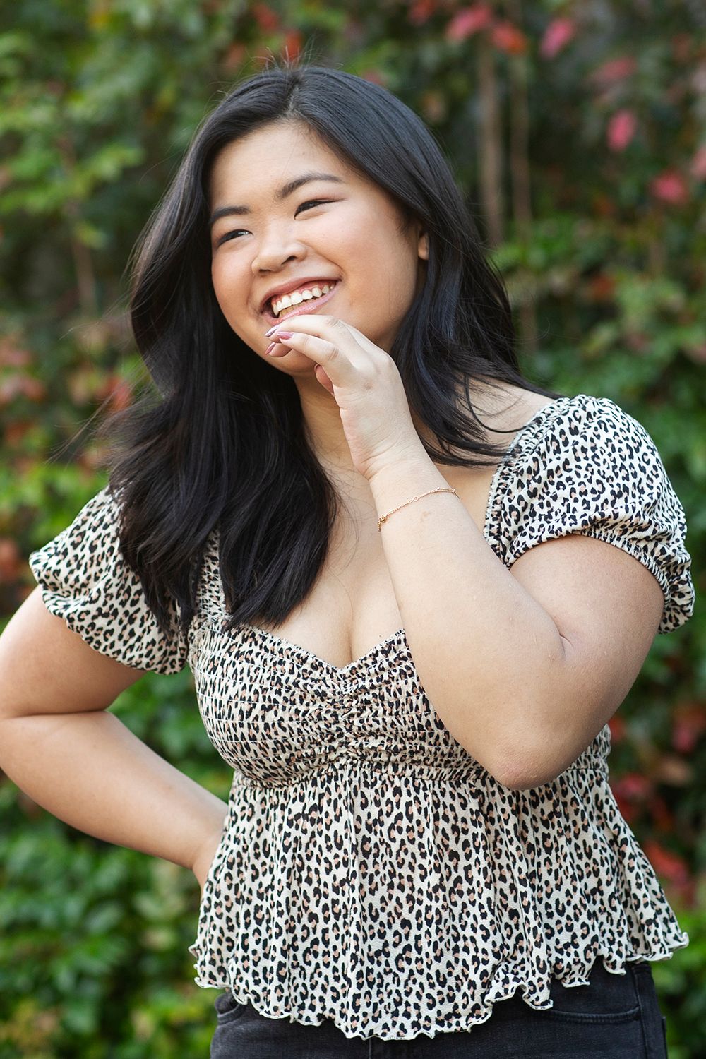 Smiling young Asian woman in leopard print top laughing outdoors with green foliage background.
