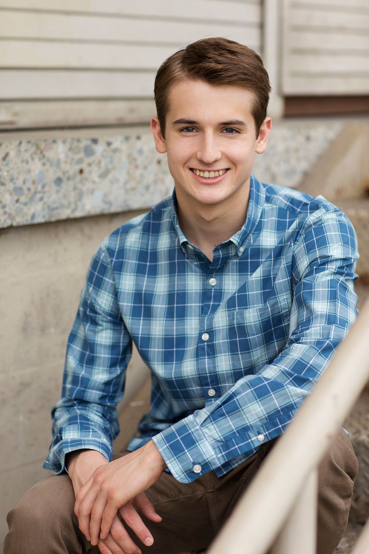Smiling young man in blue plaid shirt sitting on steps, casual portrait photo.