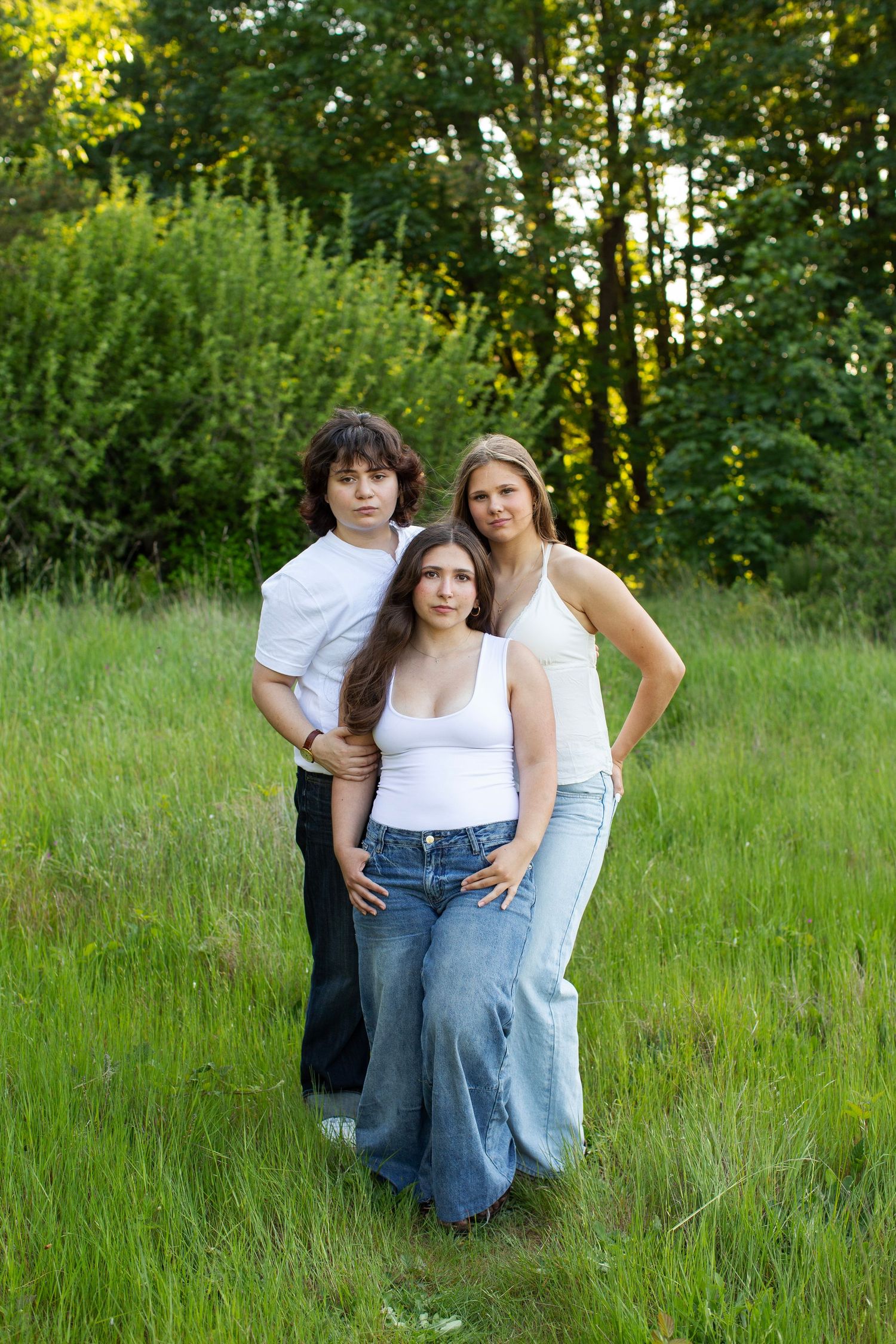 Three teenagers in white tops and jeans pose together in a lush green meadow surrounded by trees.