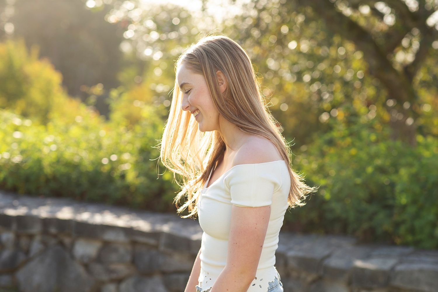 Young blonde woman in white off-shoulder top poses outdoors in golden sunlight with lush green garden backdrop.