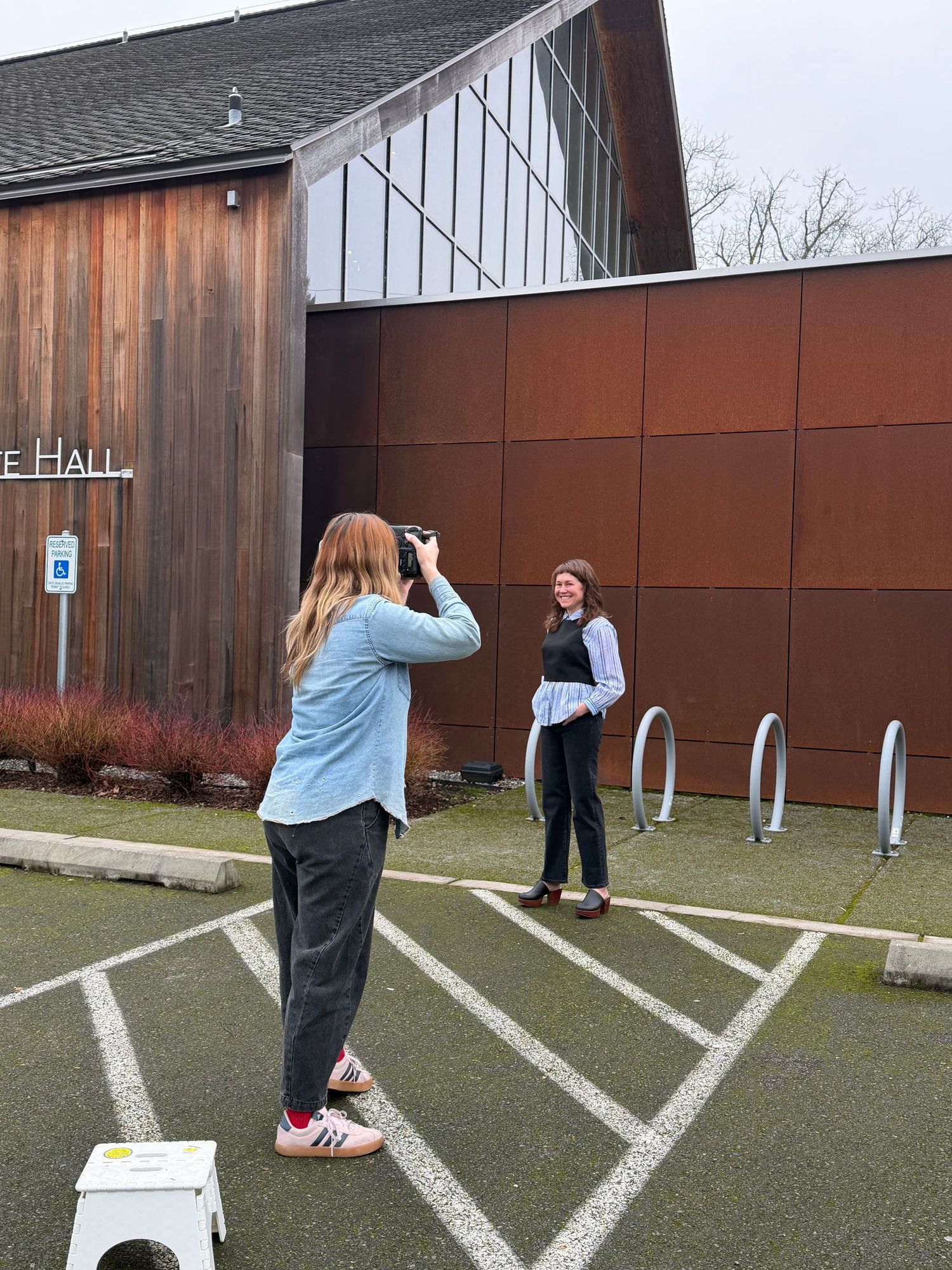 Photographer capturing portrait of woman outside modern wooden building in parking lot.