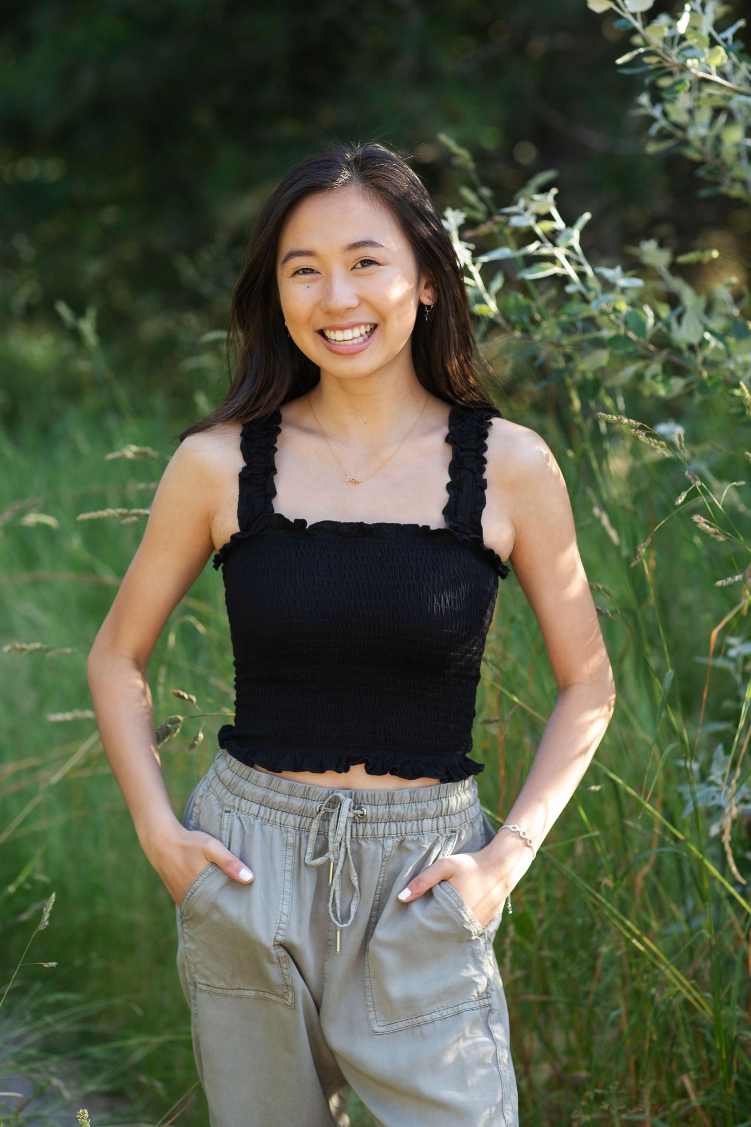 Smiling young woman in black crop top and khaki joggers posing outdoors among lush green foliage.
