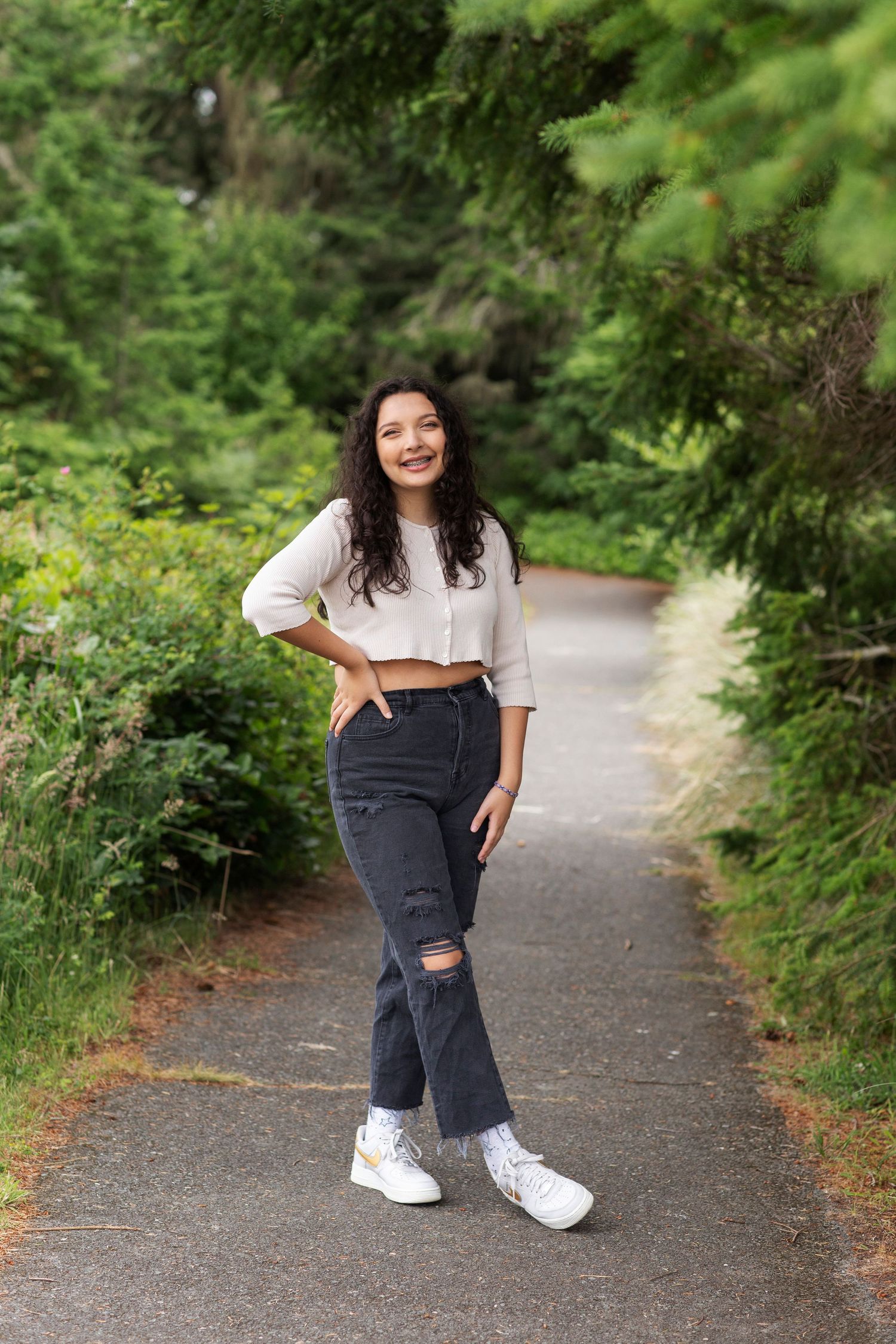 Young woman in white crop top and ripped jeans smiling on a tree-lined garden path.