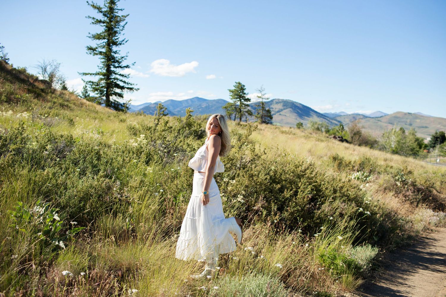Woman in white midi dress walking along a dirt path through a lush mountain meadow landscape.