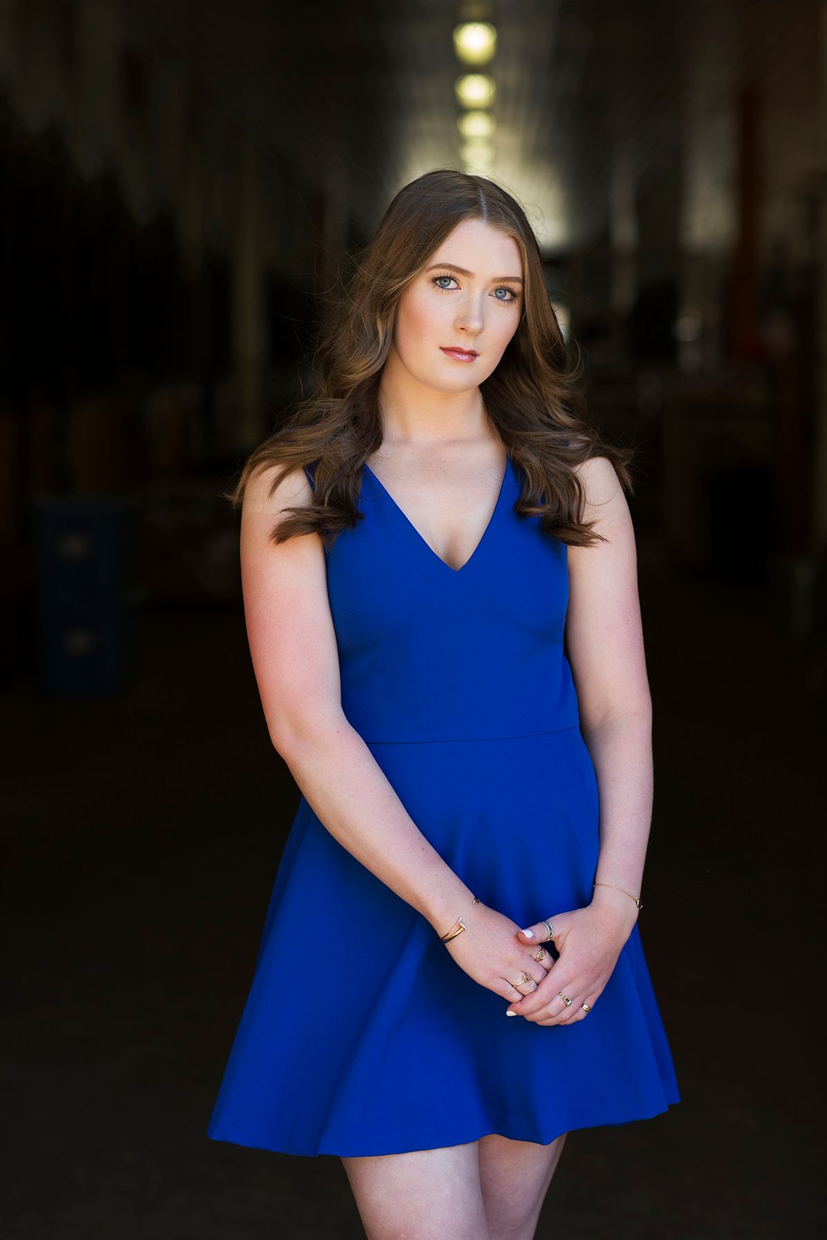 Young woman in a royal blue v-neck mini dress poses confidently in a dimly lit hallway.