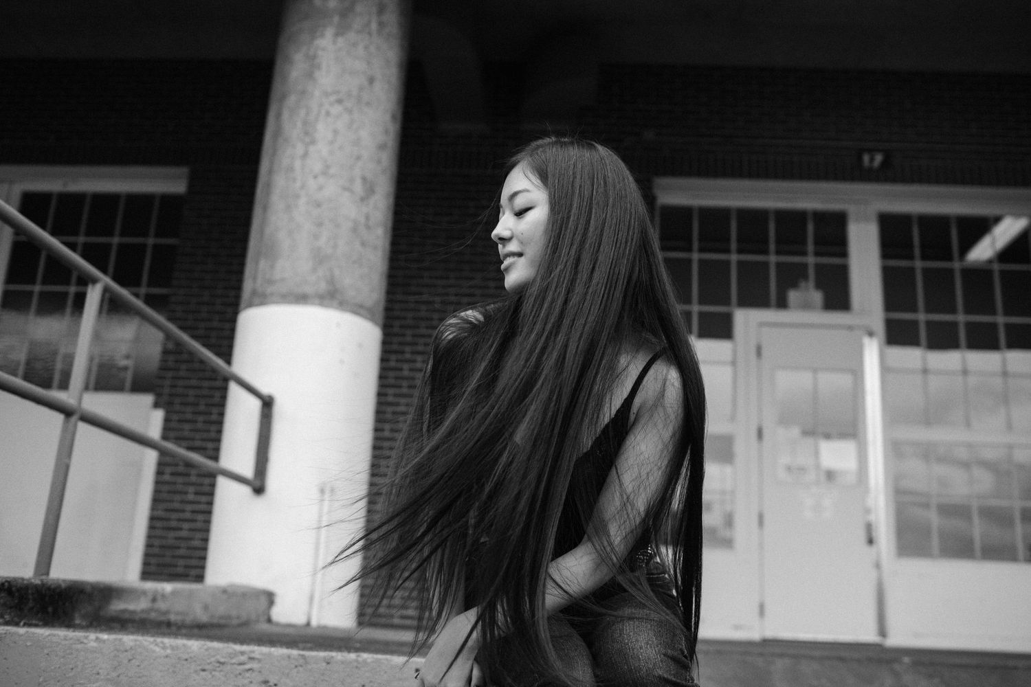 Young woman with long flowing dark hair walking outside brick building in black and white photo.