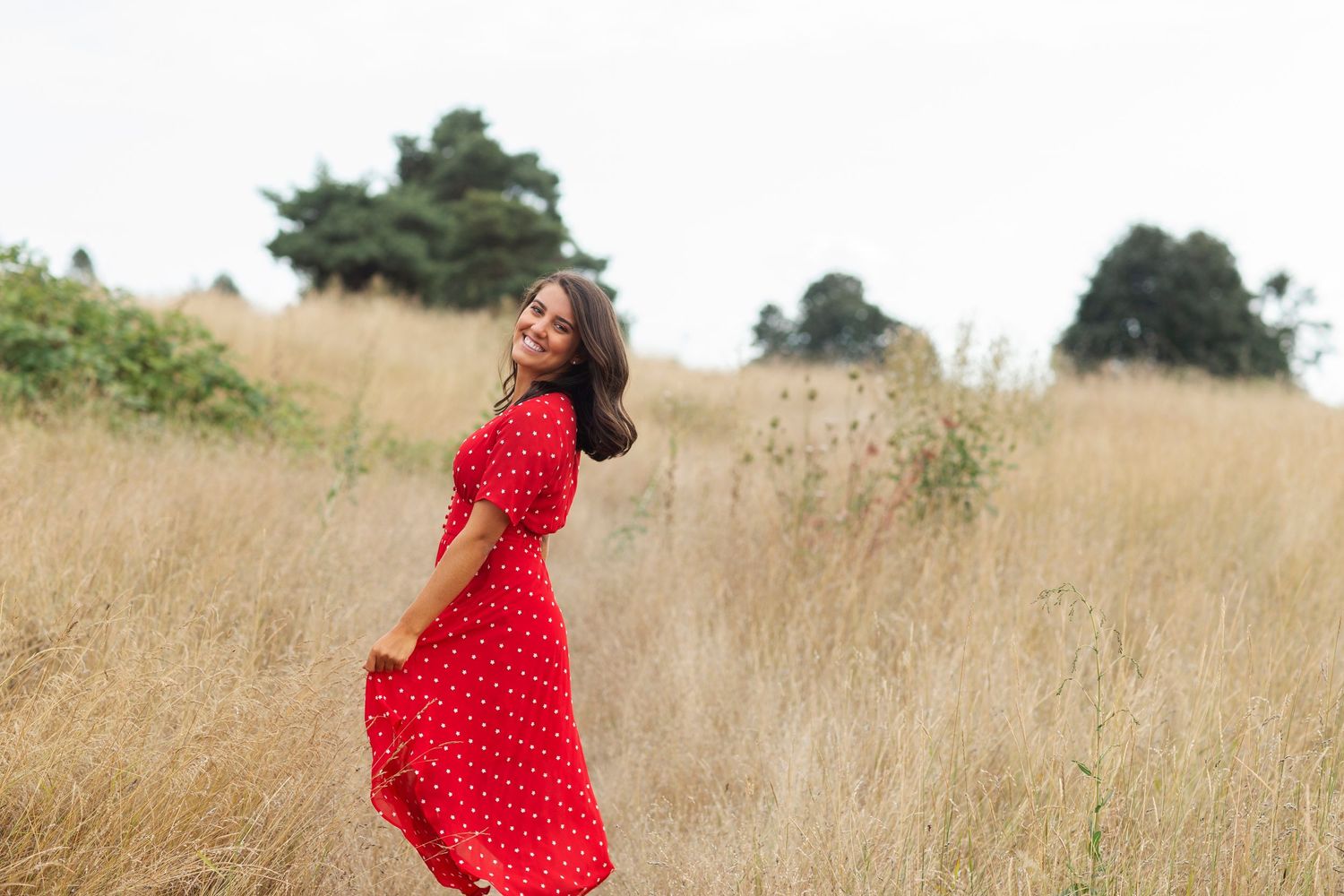 Woman in red polka dot dress twirling in golden field with trees in background.