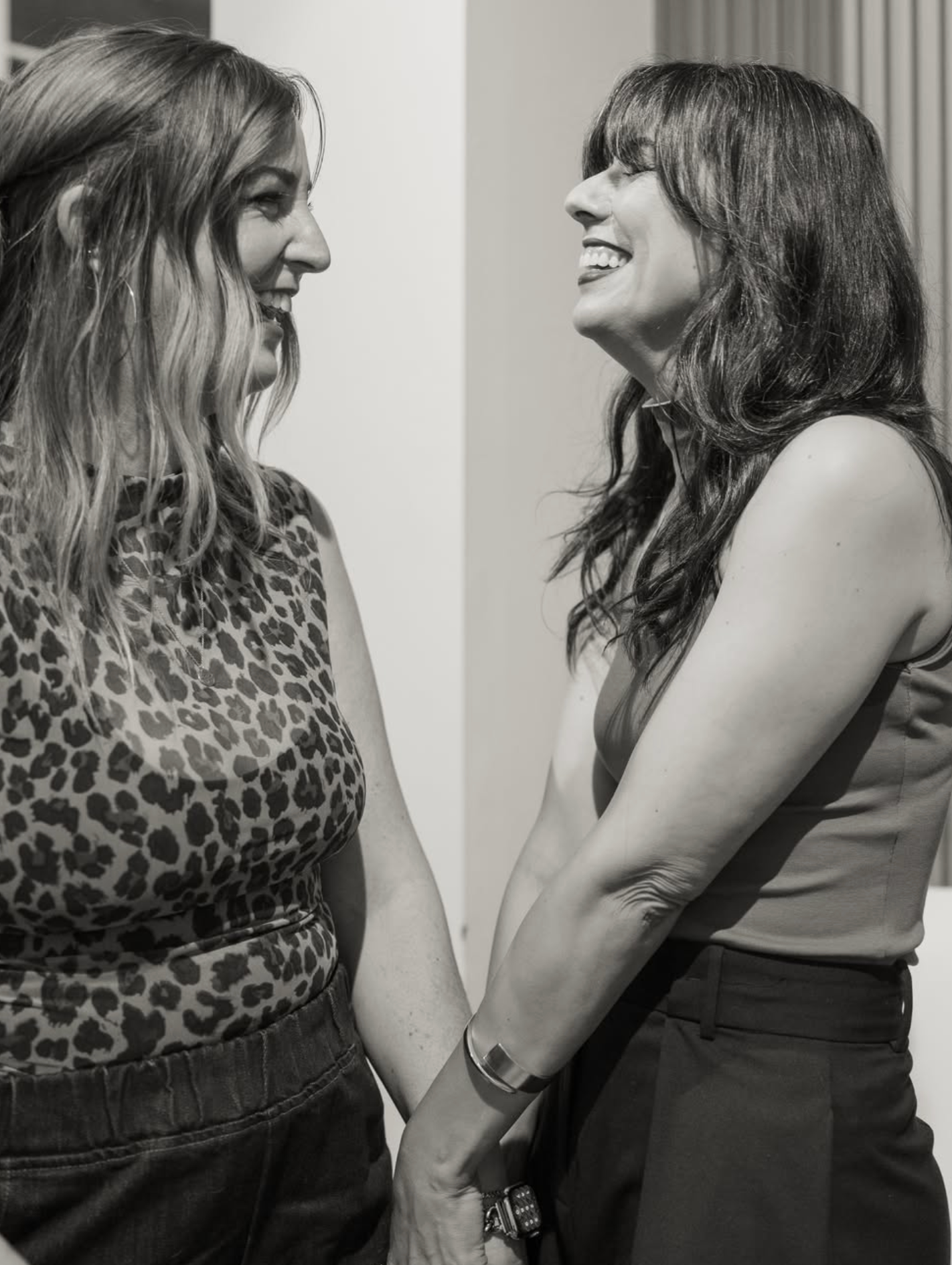 Black and white photo of two women holding hands and smiling at each other at an indoor event.