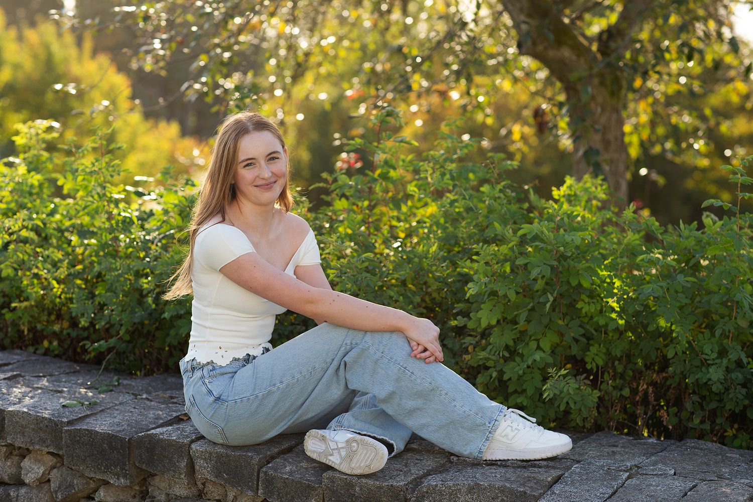 Young woman in jeans and white top sitting on stone wall surrounded by lush green garden in golden sunlight.