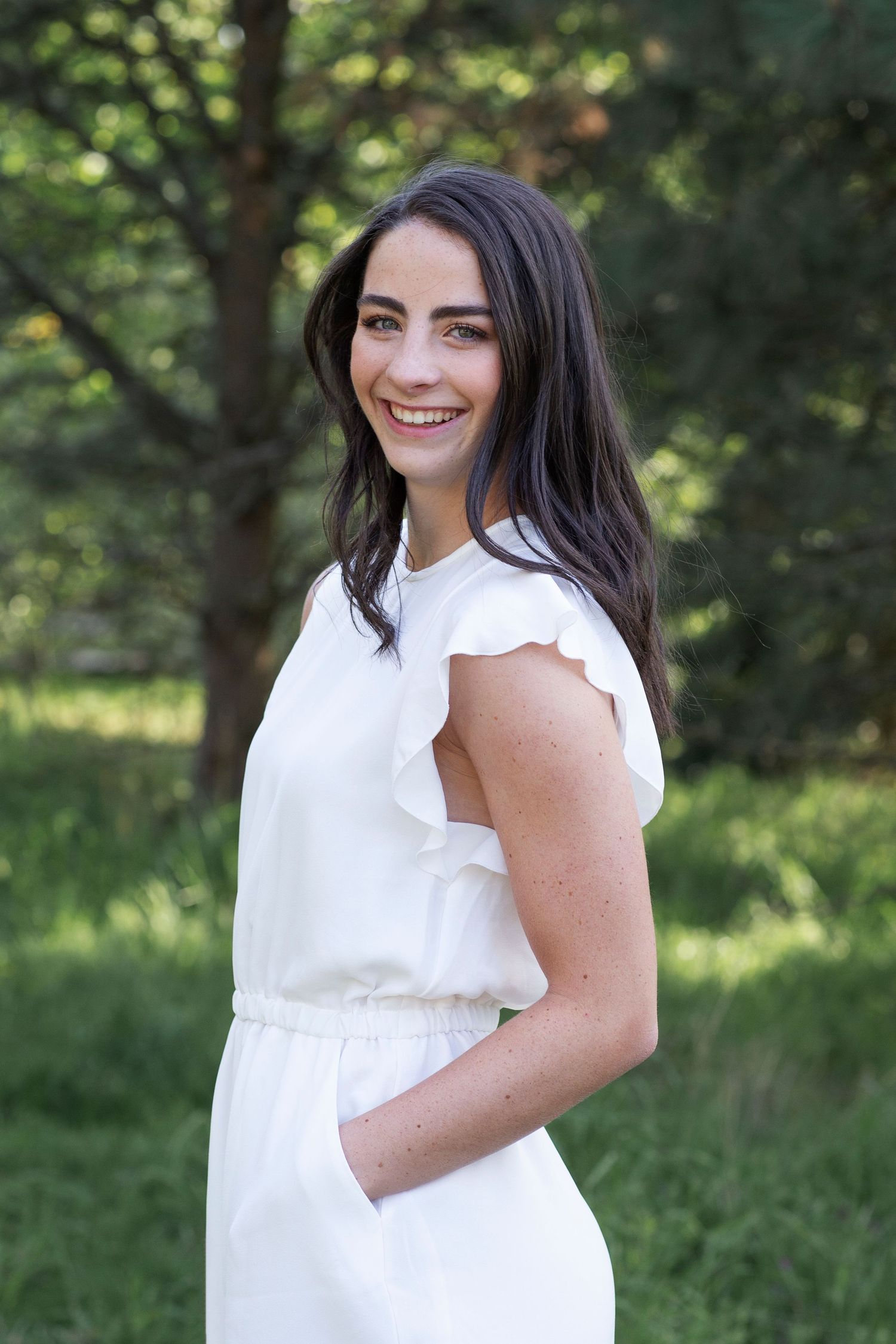 Smiling young woman with dark hair wearing a white ruffled dress, standing outdoors among green trees.